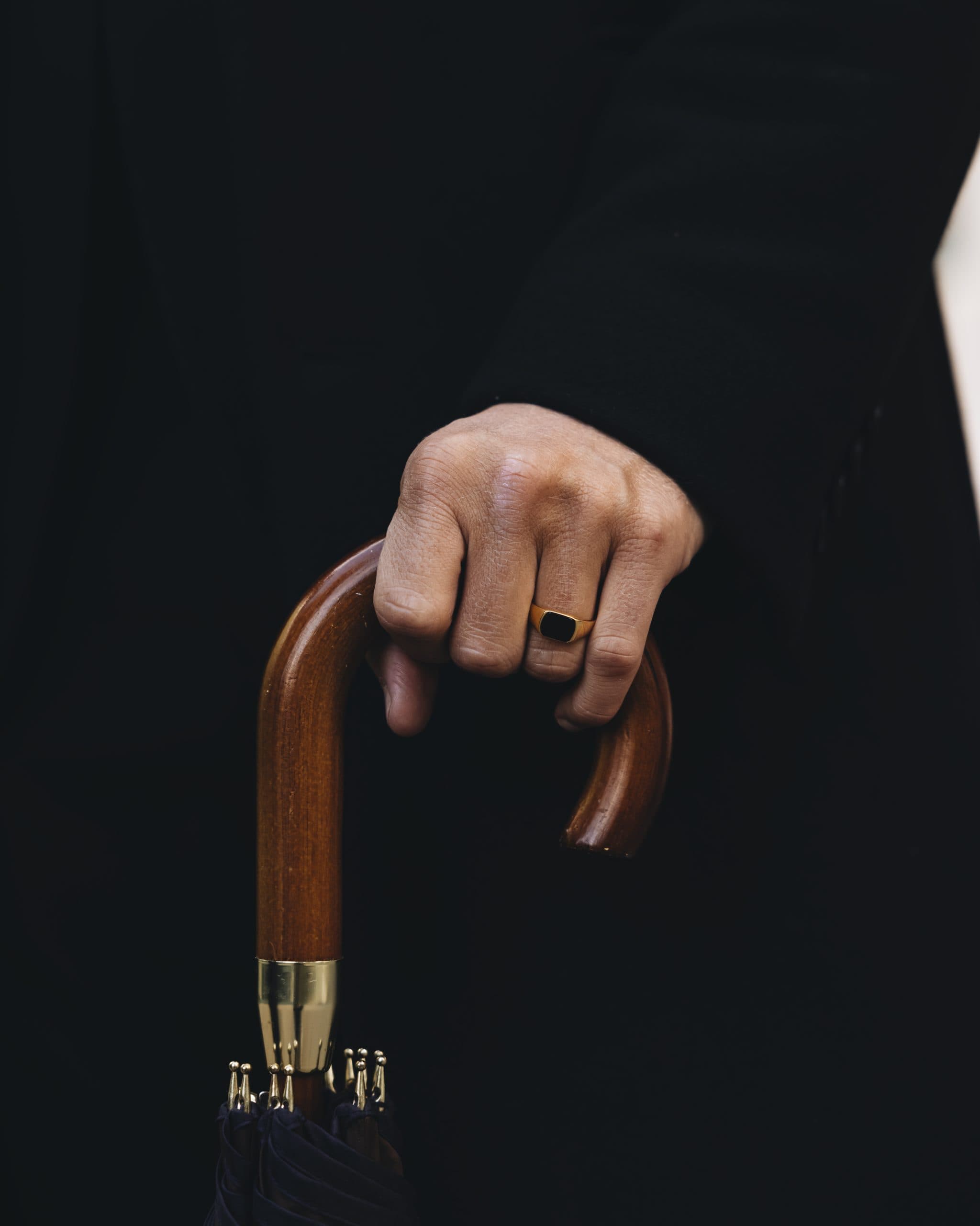 A hand resting on a wooden walking cane, gold signet ring, dark background
